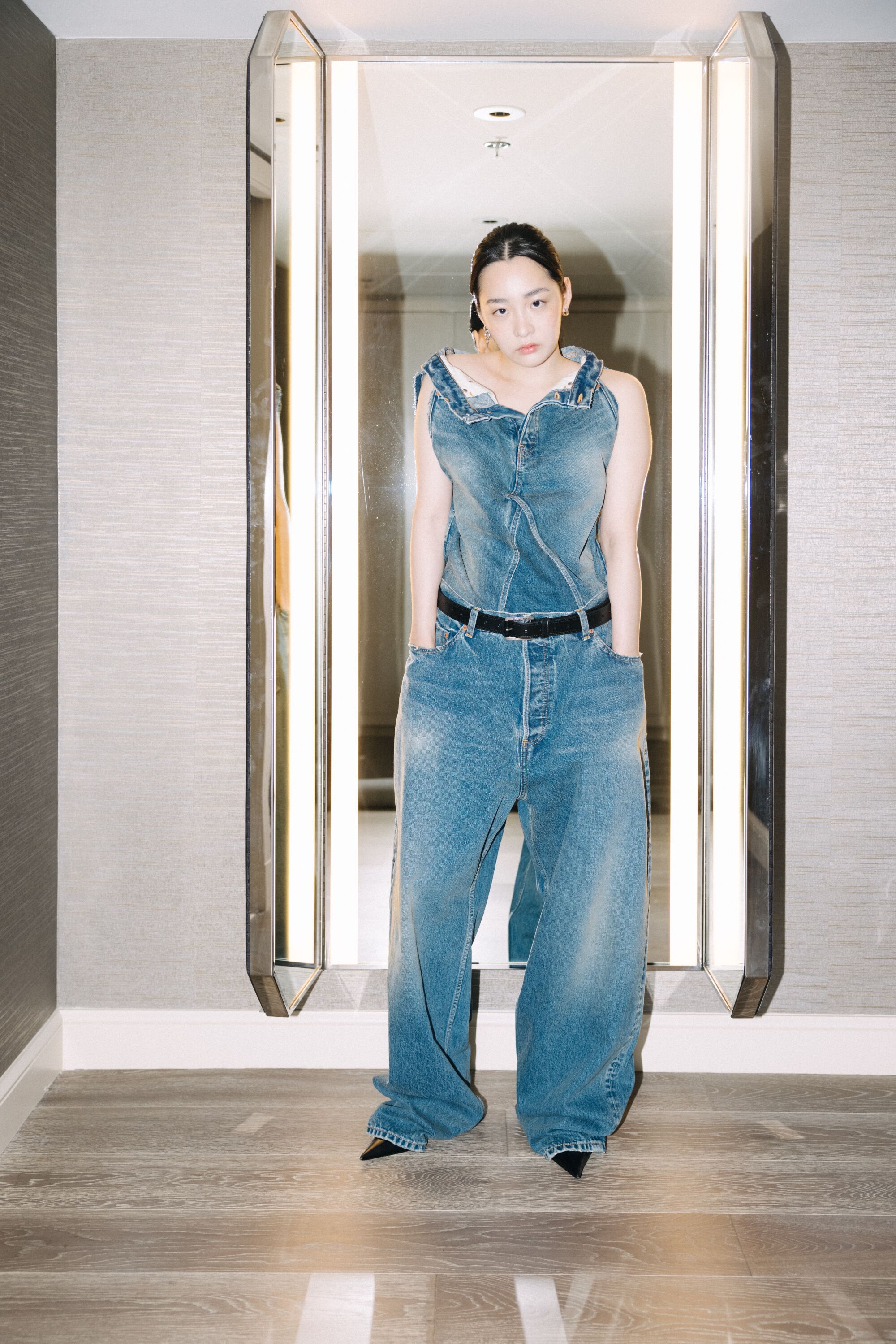 A woman in a faded denim outfit poses for a photo against a wall mirror.