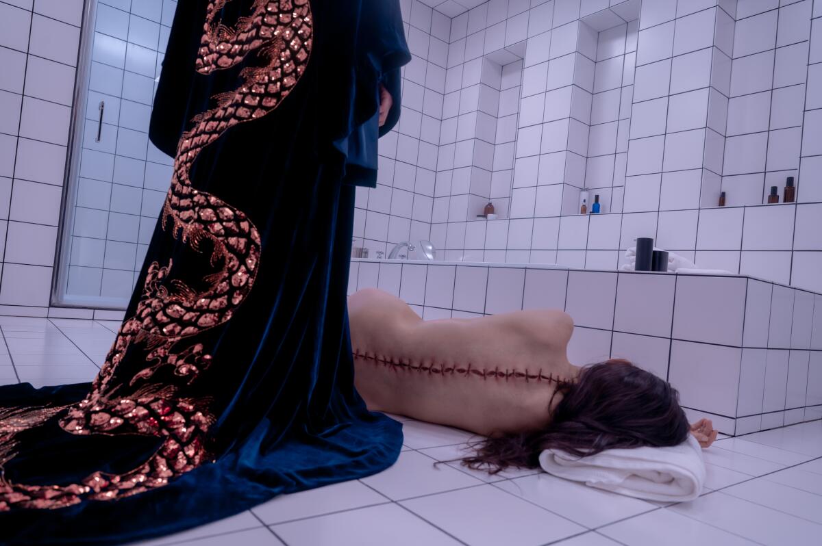 A woman in a robe stands over a woman with a sewn-up back lying on the floor.