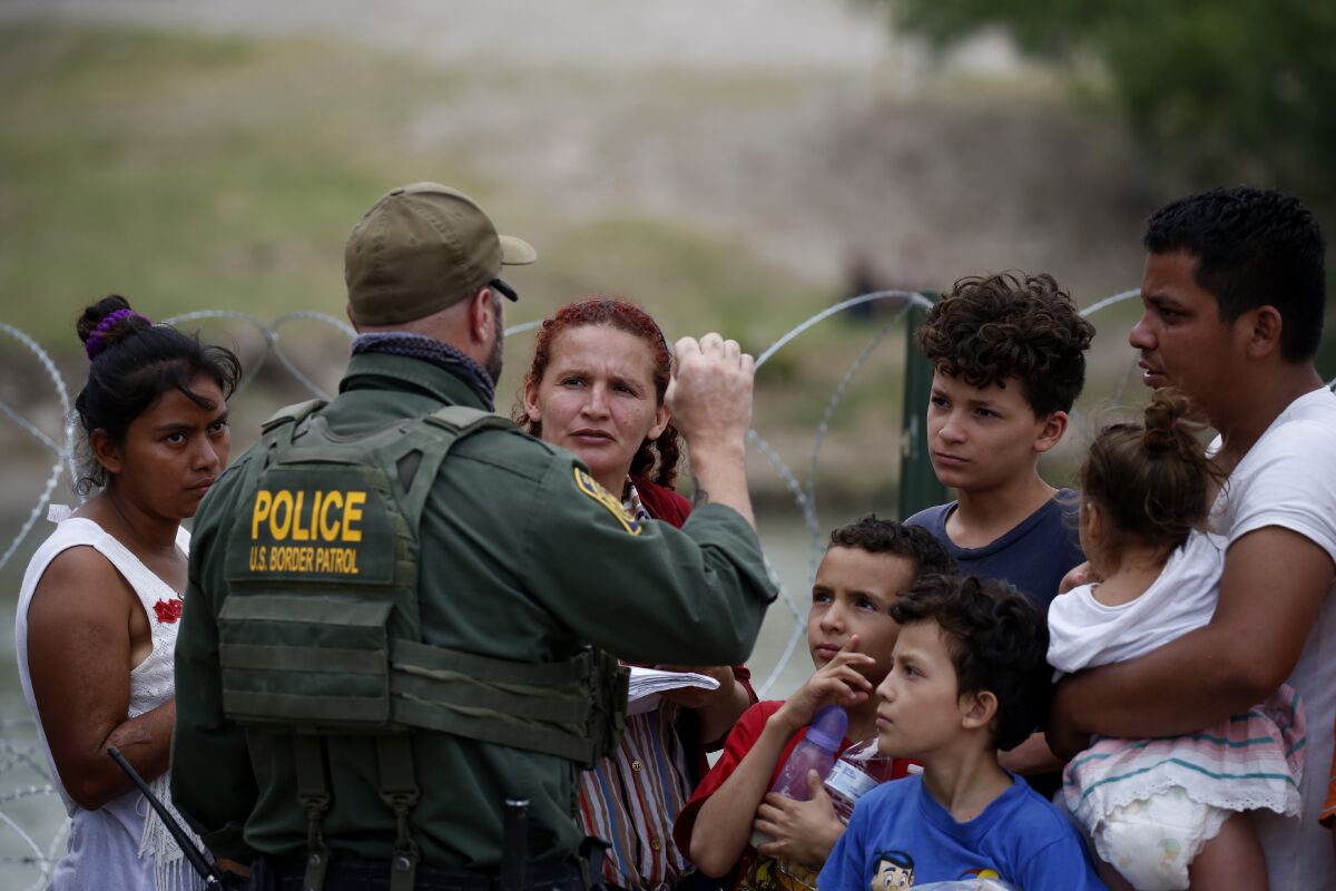 A man in a green U.S. Border Patrol police uniform speaking to a small group of men, women and children outside.