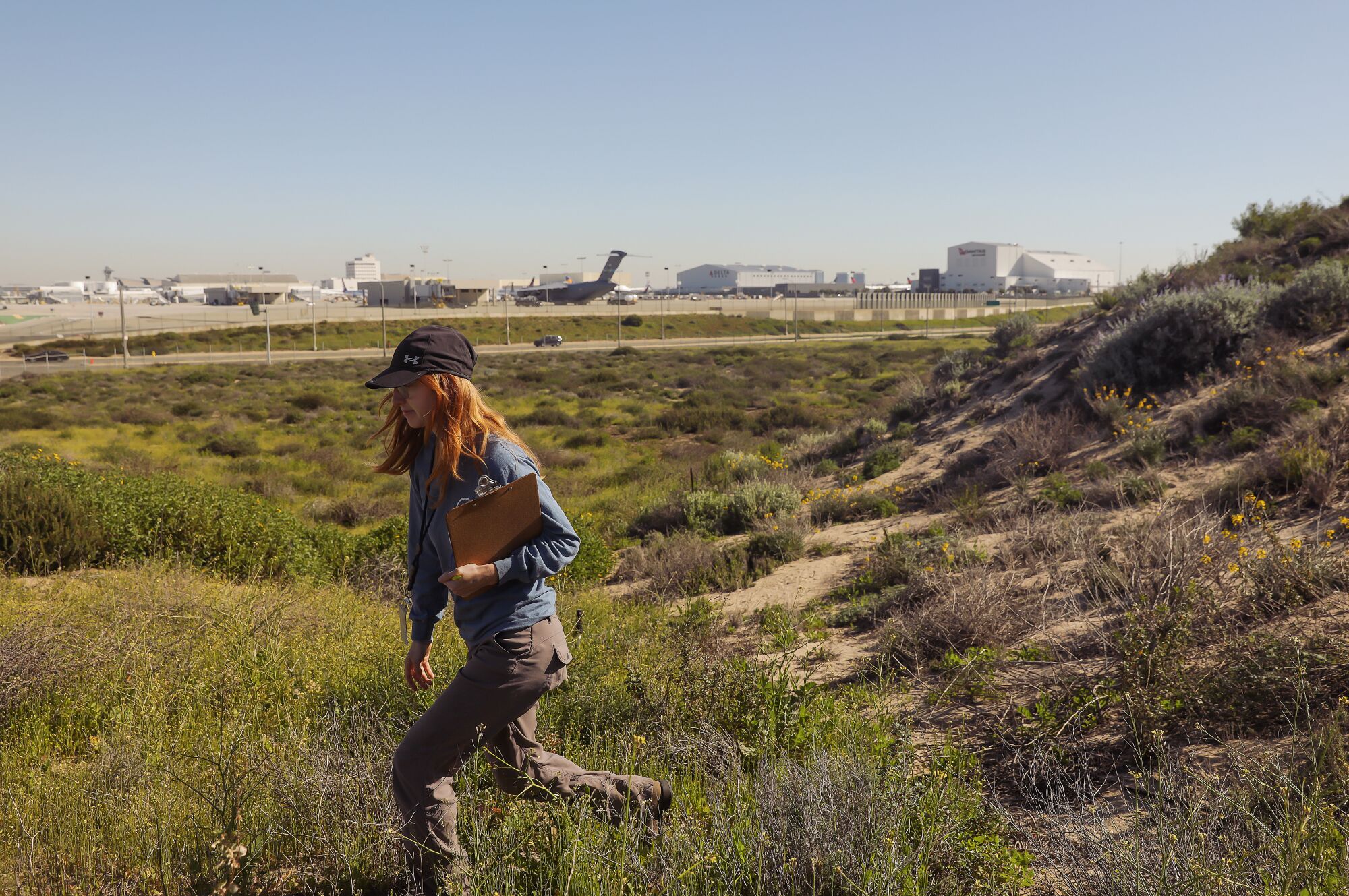 A woman wearing a baseball cap and holding a clipboard walks through greenery, with parked airplanes in the background.