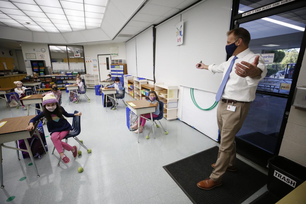 A school administrator stretches his arms out at the door of a classroom of young children at spaced-out desks