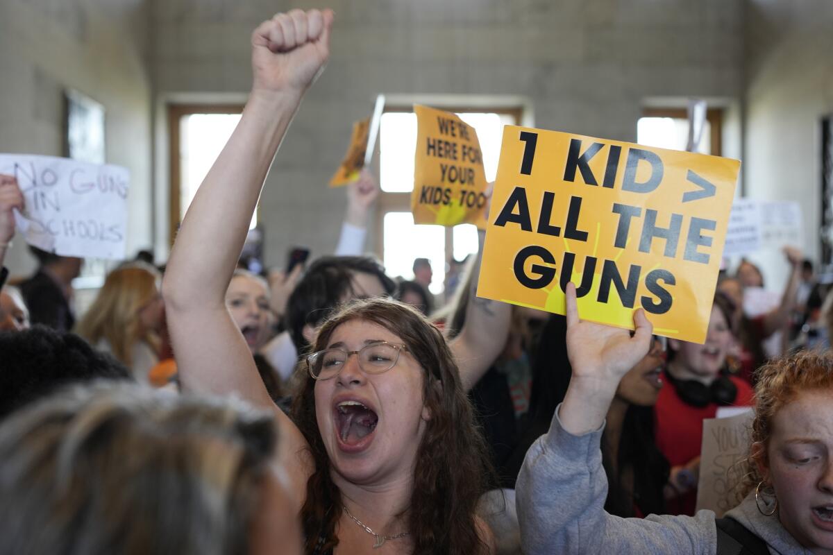 A crowd of protesters indoors, one raising a fist and others waving signs with messages including, "1 kid > all the guns."