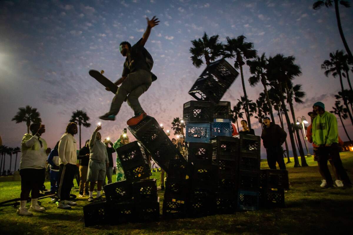 People stand around at dusk watching a man fall off a large pyramid of milk crates