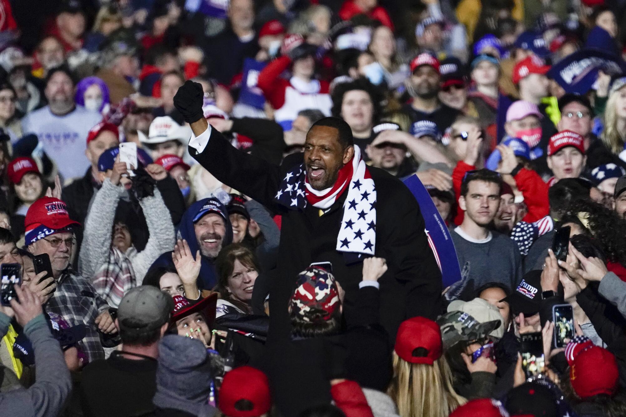Amid a crowd of people in red hats, a man in a winter scarf with a U.S. flag motif speaks.
