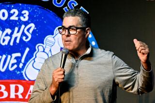 Adam Carolla speaking into a microphone and lifting one hand while clad in a casual long-sleeve gray shirt