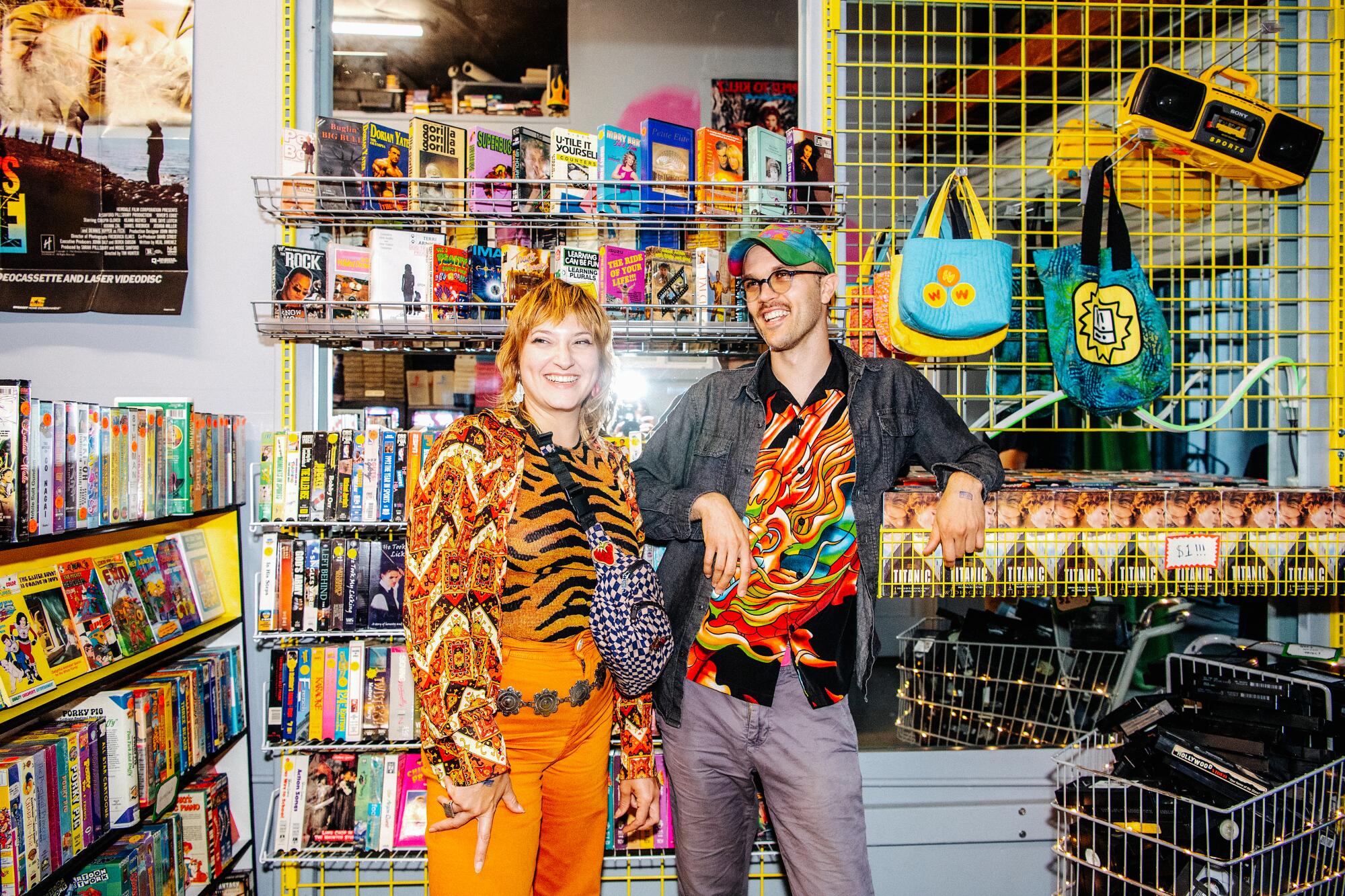 A woman, left, and a man in bright outfits smile among shelves of VHS tapes 