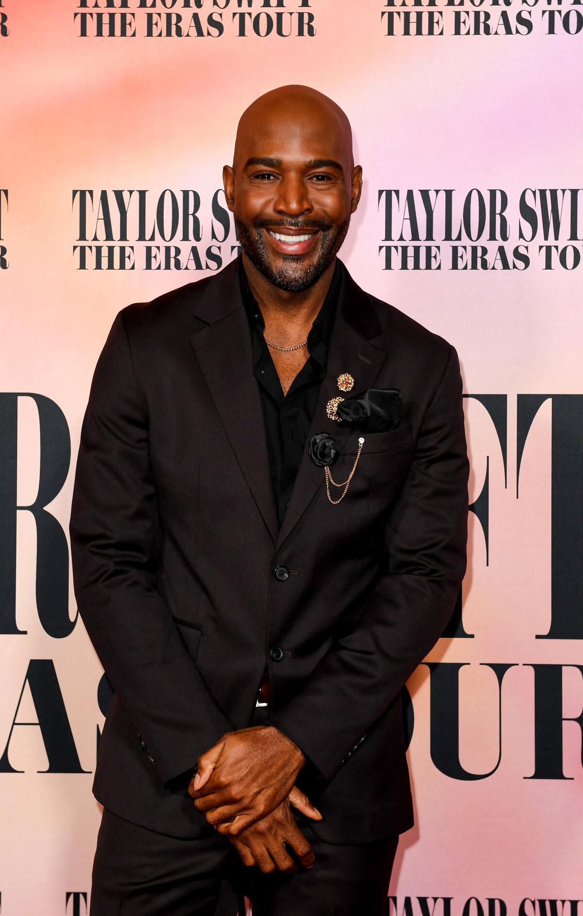 Karamo Brown, in a black suit and open-collar shirt, stands with his hands crossed in front of him