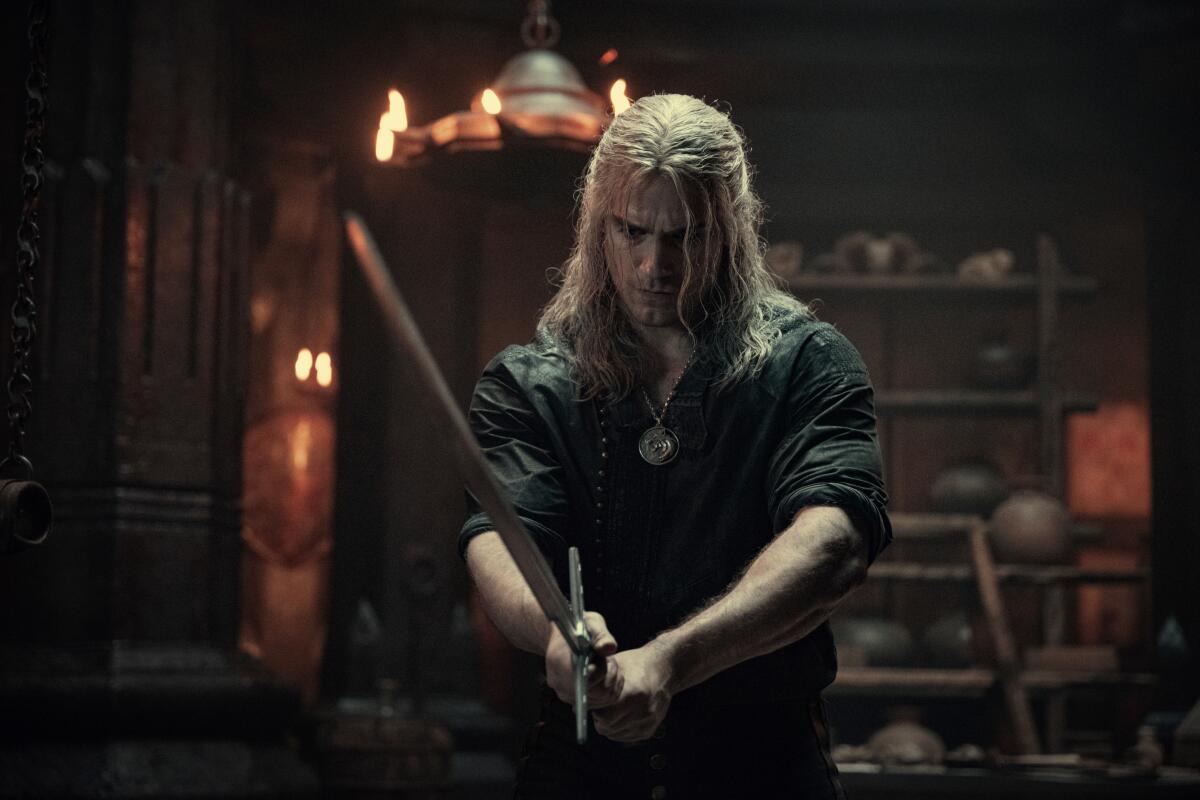 Henry Cavill, with long blond hair that partially covers his face, holds up a sword in "The Witcher."
