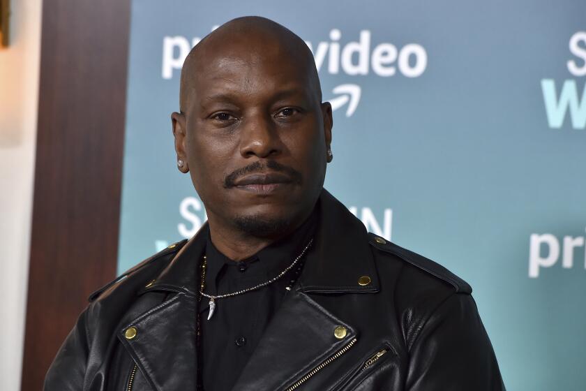 Tyrese Gibson in a black shirt, a chain and black leather jacket posing for pictures against a blue backdrop
