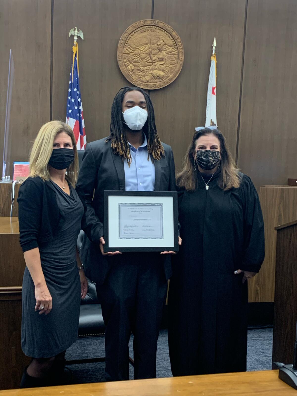Three people pose in a courtroom with a framed certificate