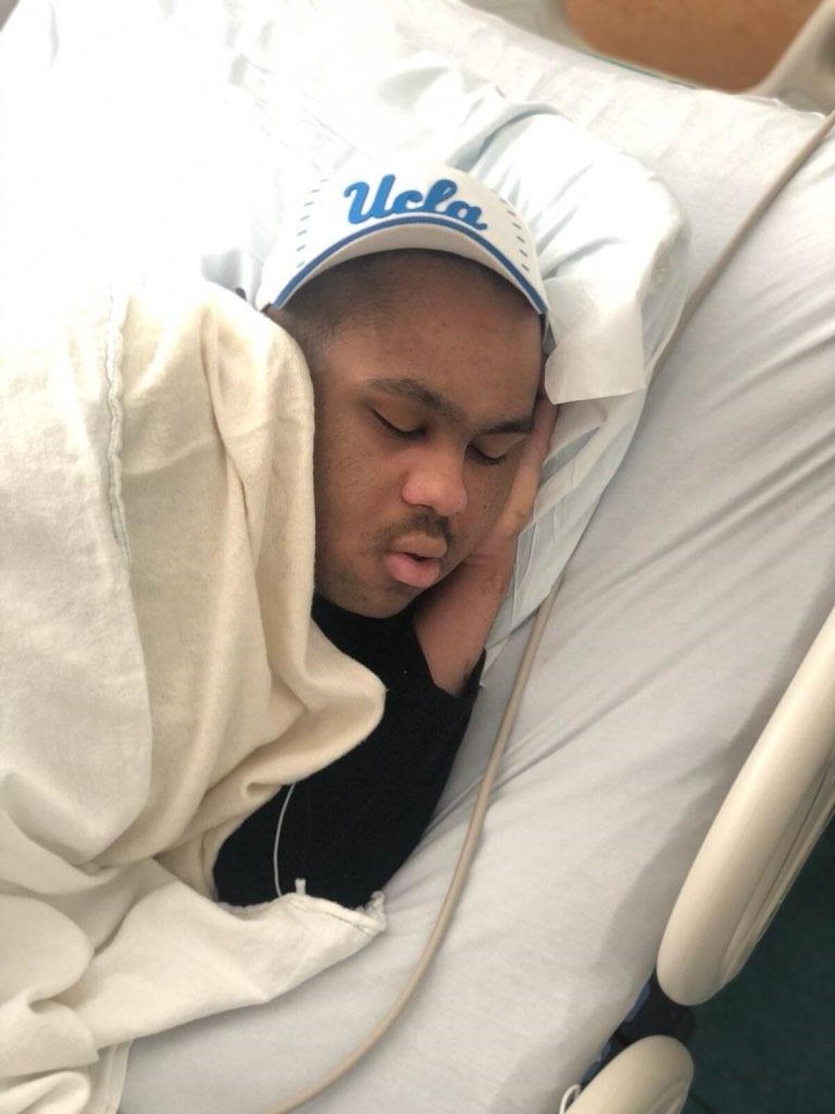Luke Forbes sleeps in a hospital bed while wearing a UCLA hat