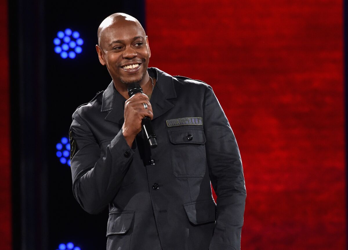 Dave Chappelle holds a microphone and smiles.