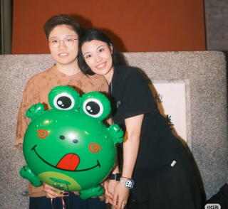 Two women holding hands pose with a balloon of a frog