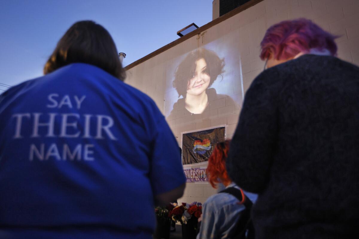 A person with a jacket reading "Say their name" stands at a vigil whre a photograph of Nex Benedict is projected on a wall. 