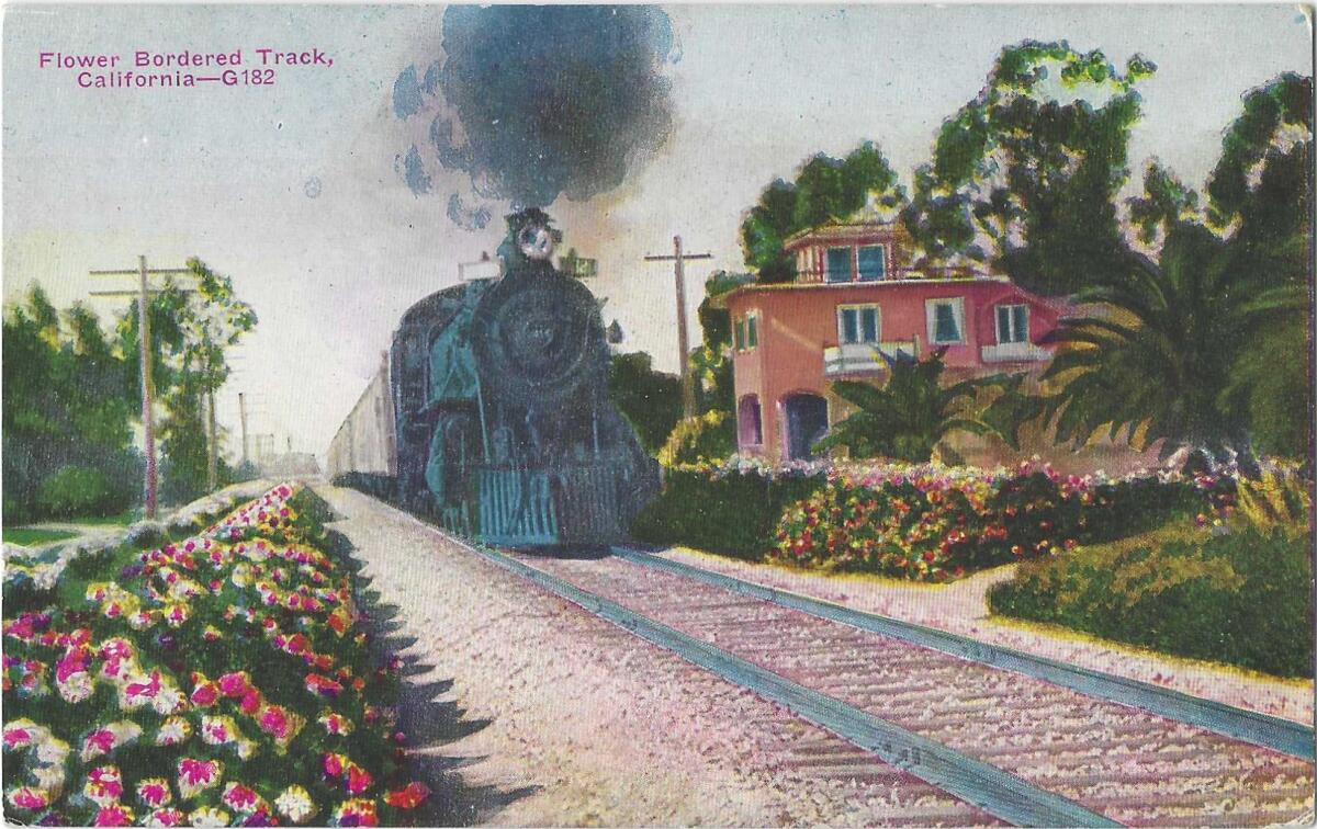 Scan of postcard shows a locomotive with billowing smoke, train tracks lined by flowers and foliage and a pretty building.