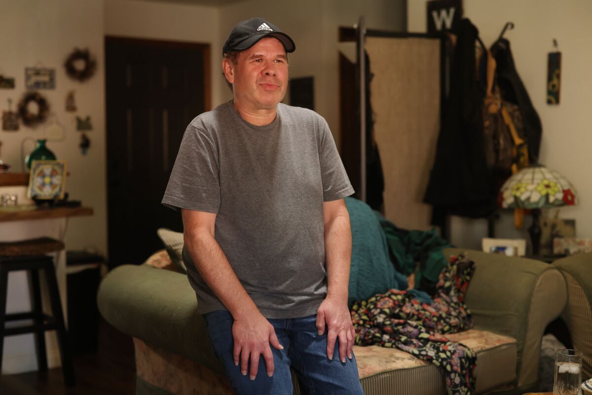 A man in a ball cap smiles while sitting on the arm of a sofa.