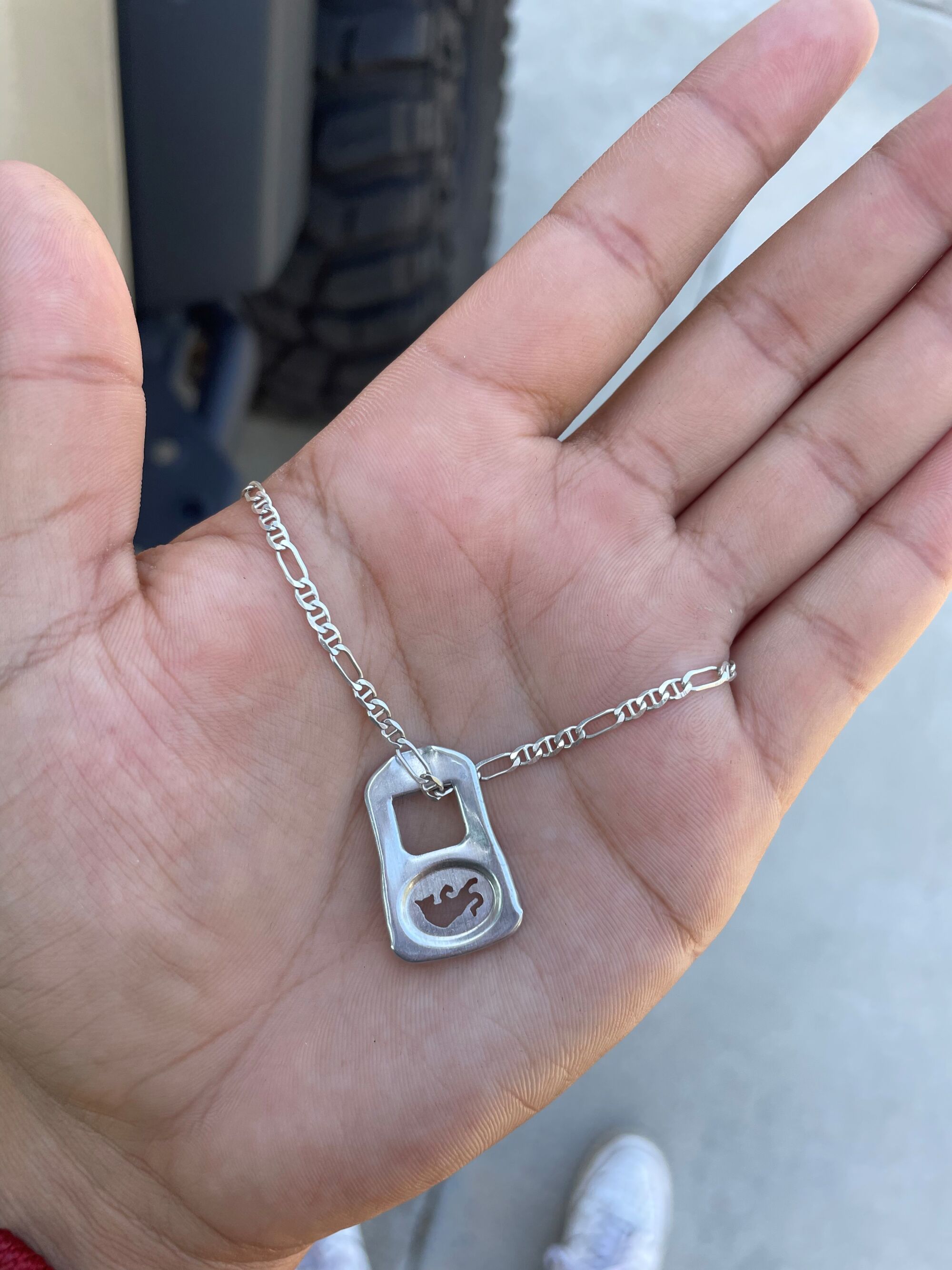 A hand holding a silver necklace with a drink can tab on it.