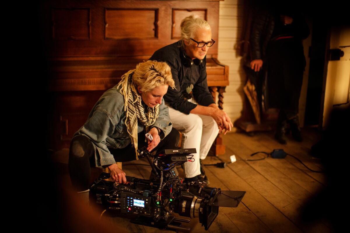 Two women sit in front of an old-fashioned upright piano, one leaning over a movie camera.