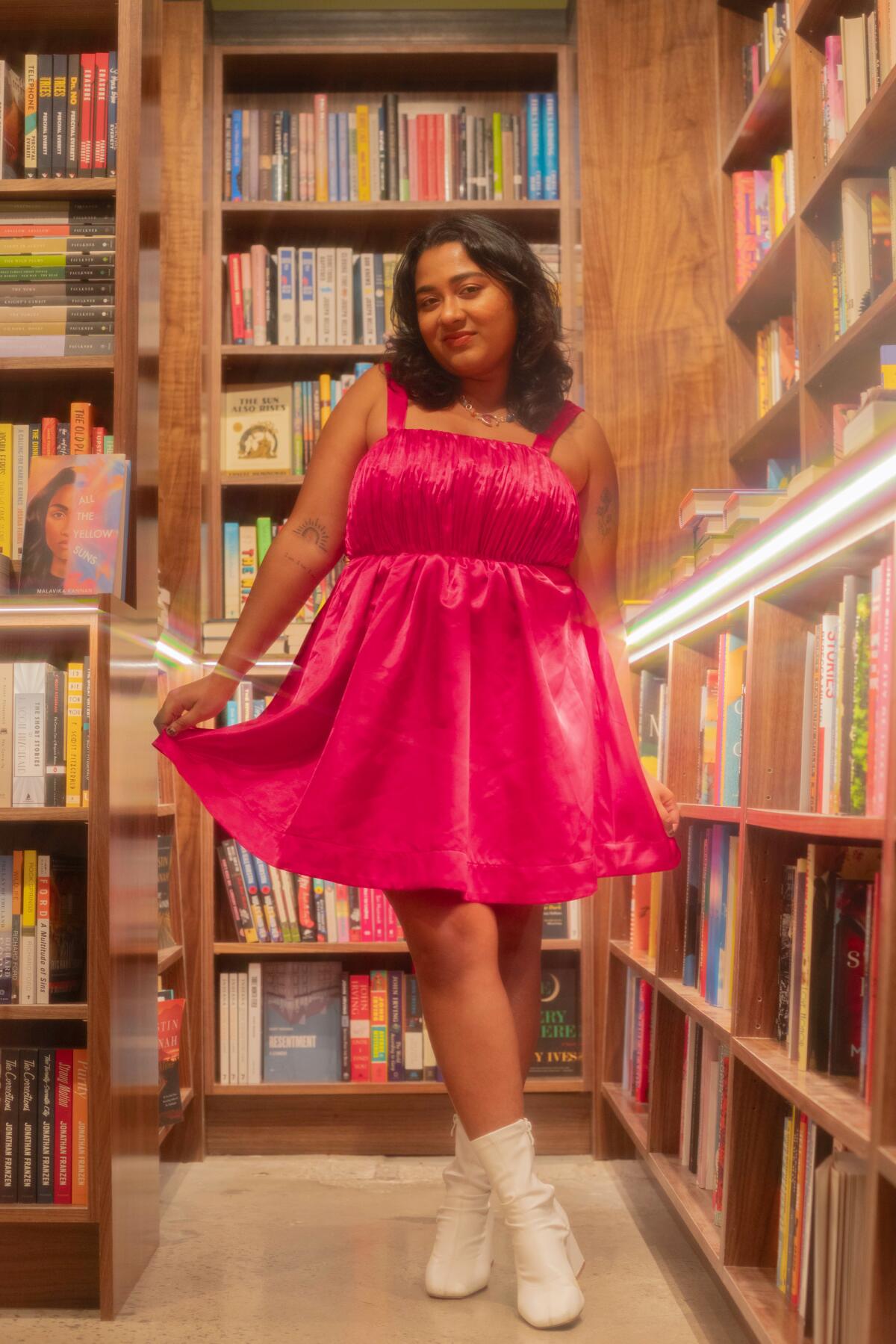 A woman in a pink dress and white boosts poses among shelves full of books.