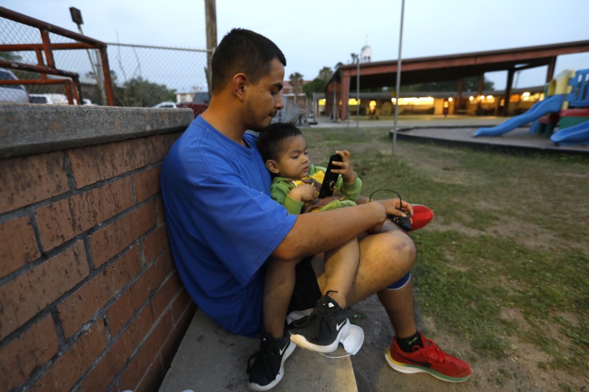A man holds his son, who is looking at a cellphone, in his lap while sitting against a brick wall