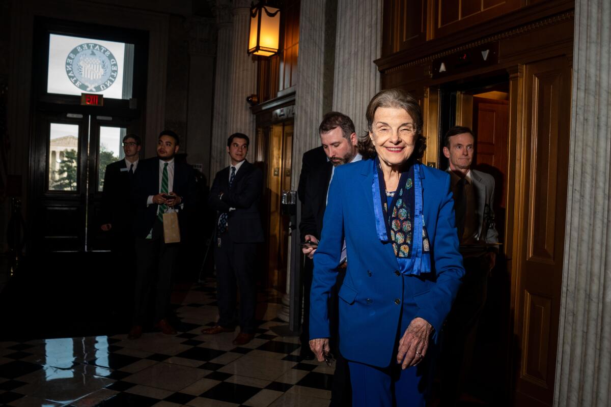 A woman in a blue jacket and pants and patterned blue scarf smiles as she walks in front of several men near a doorway