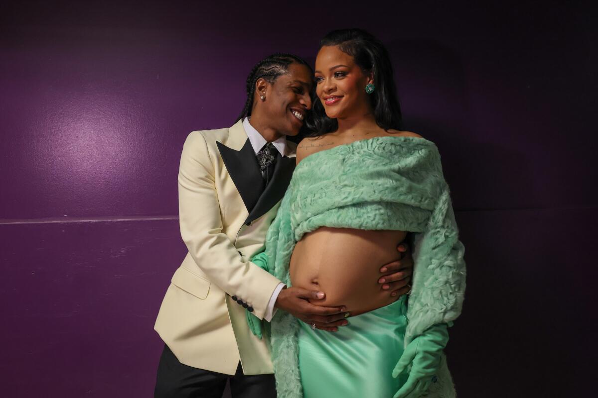 A man in a tuxedo holds his hand to the belly of a pregnant woman in a sleek dress.