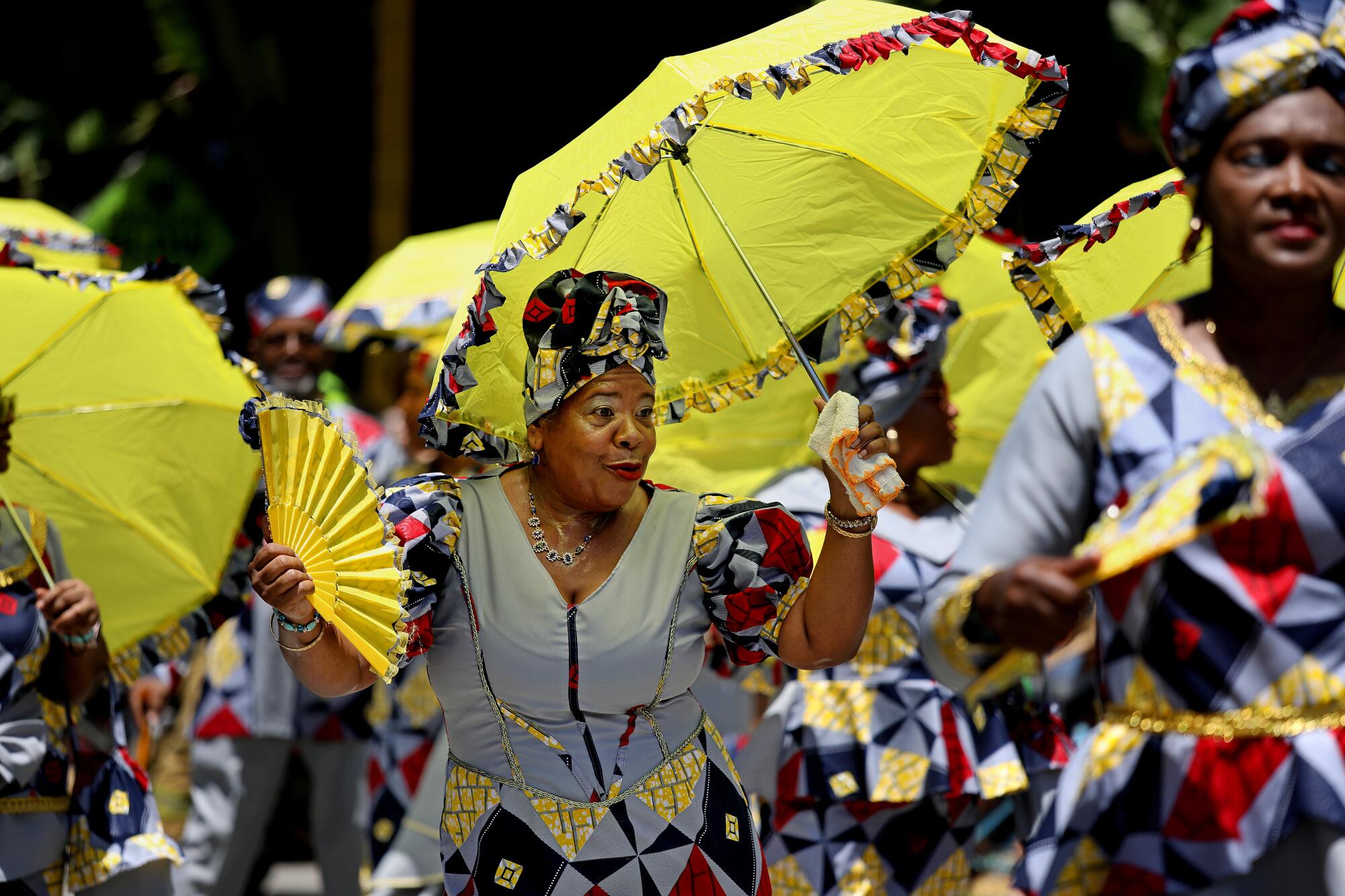 Women dance in a parade, holding yellow umbrellas and fans.