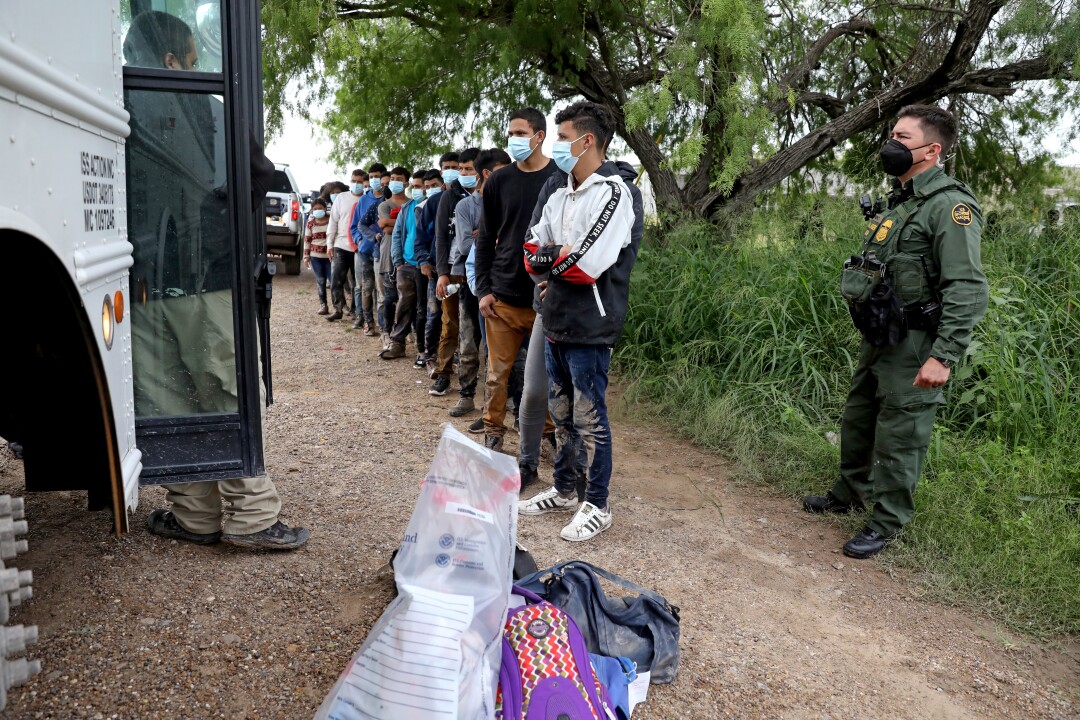 یک مامور گشت مرزی در سمت راست در نزدیکی صف مهاجرانی ایستاده است که منتظر یک اتوبوس هستند
