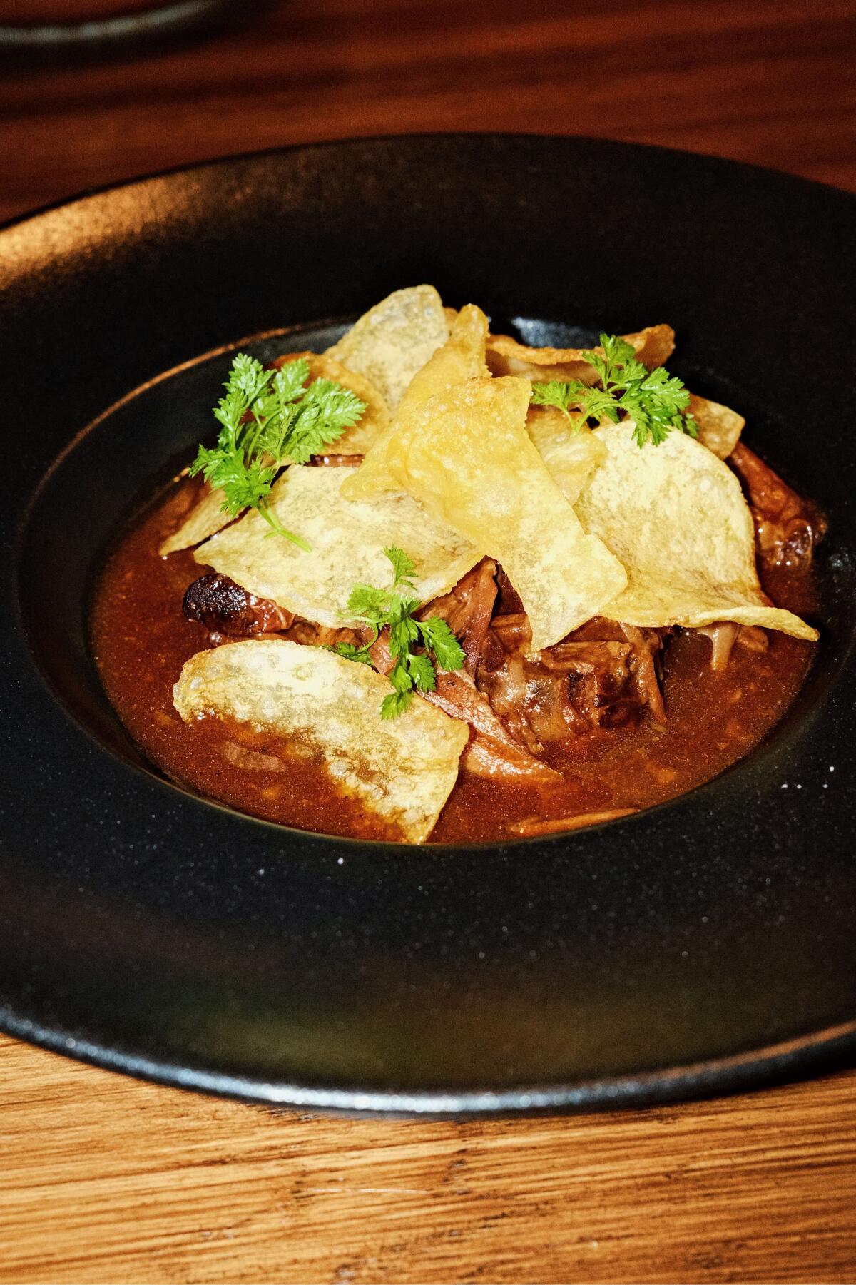 Oxtails simmered in sauce topped with chips and herbs in a black wide-brimmed bowl at Spanish restaurant Casa Leo