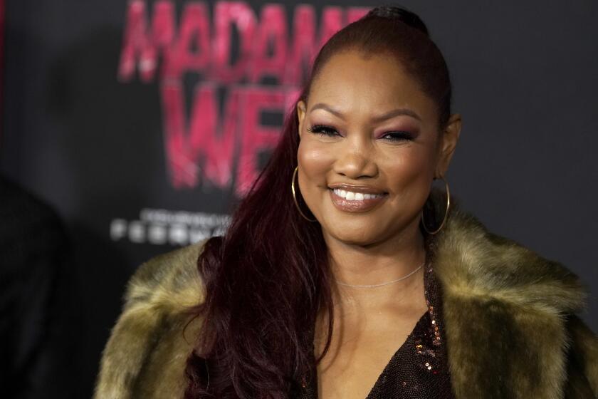 Garcelle Beauvais wearing a fur coat and hoop earrings smiling and posing against a black background with red text