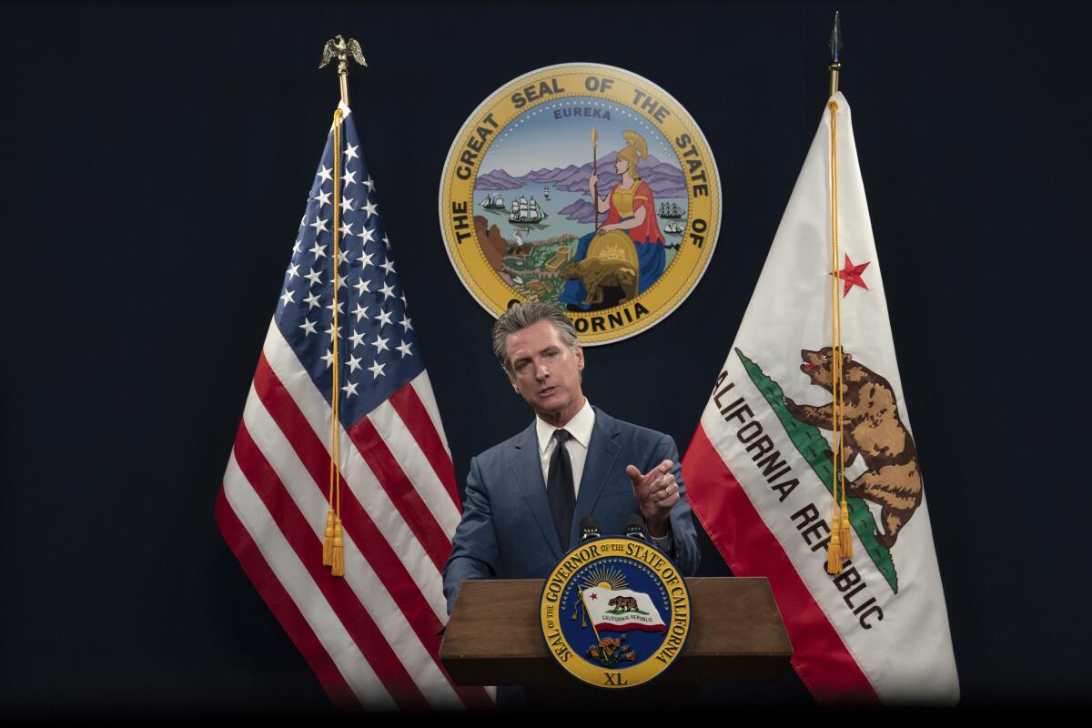 A man in suit and tie, flanked by U.S. and California flags, speaks at a lectern with an official seal behind him 