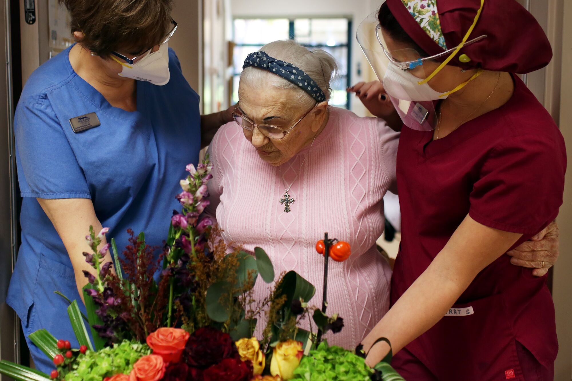 In front of an elaborate flower arrangement, two women in scrubs and masks stand with their arms around an older woman. 