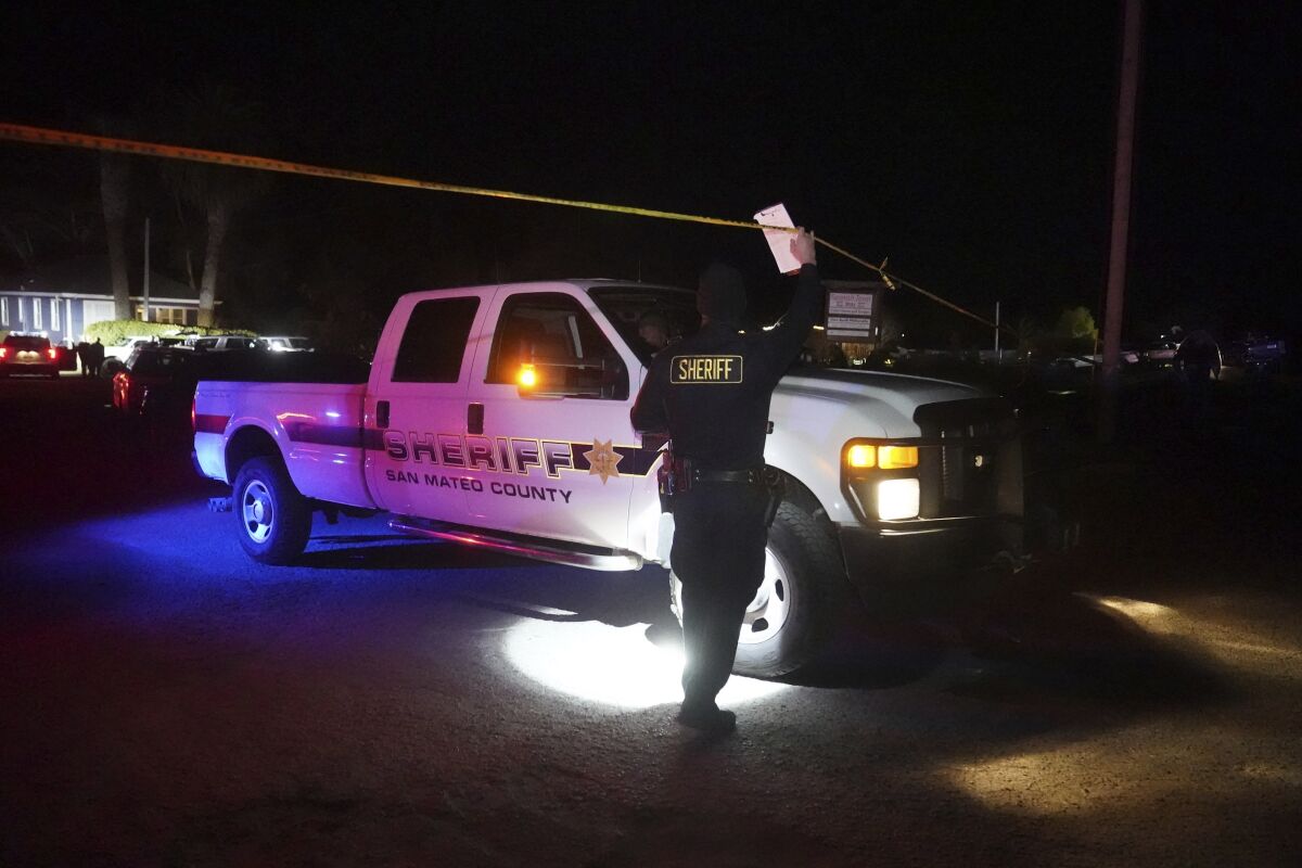 A sheriff's official, using a flashlight in the dark, lifts police tape as a San Mateo County Sheriff's truck drives under it