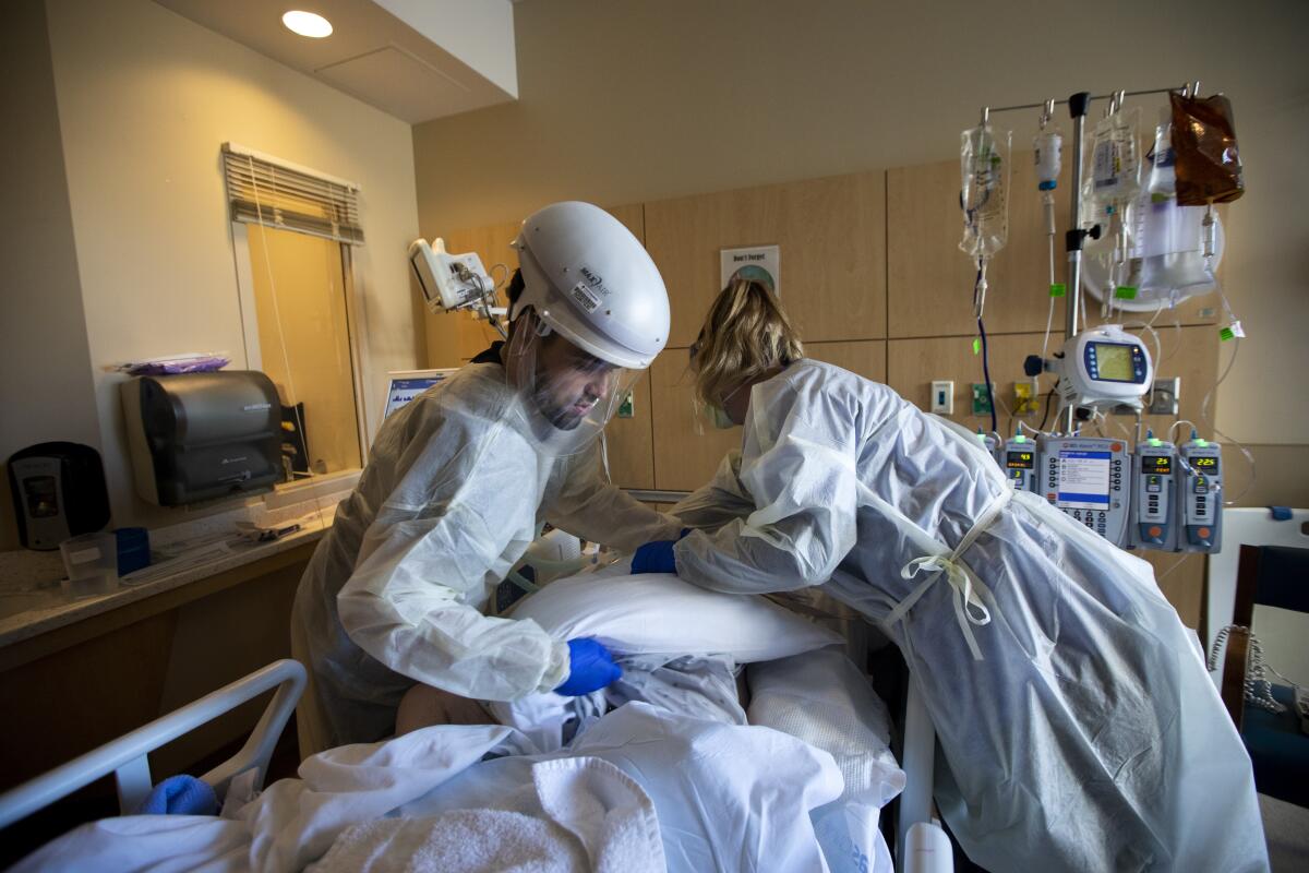Two nurses in protective gear adjust pillows and move a patient in a hospital bed
