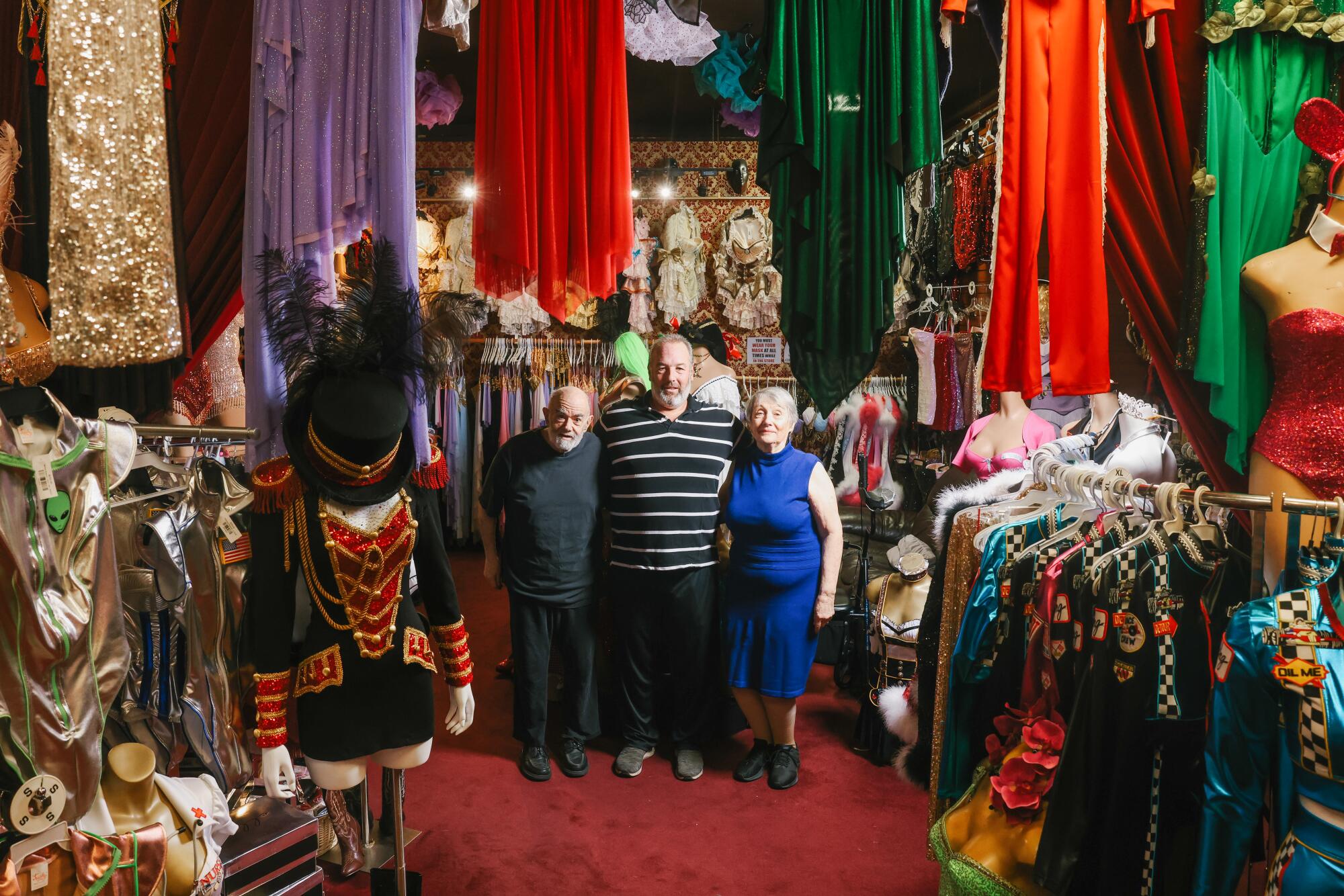 A man stands between his parents, surrounded by costumed mannequins and other items.