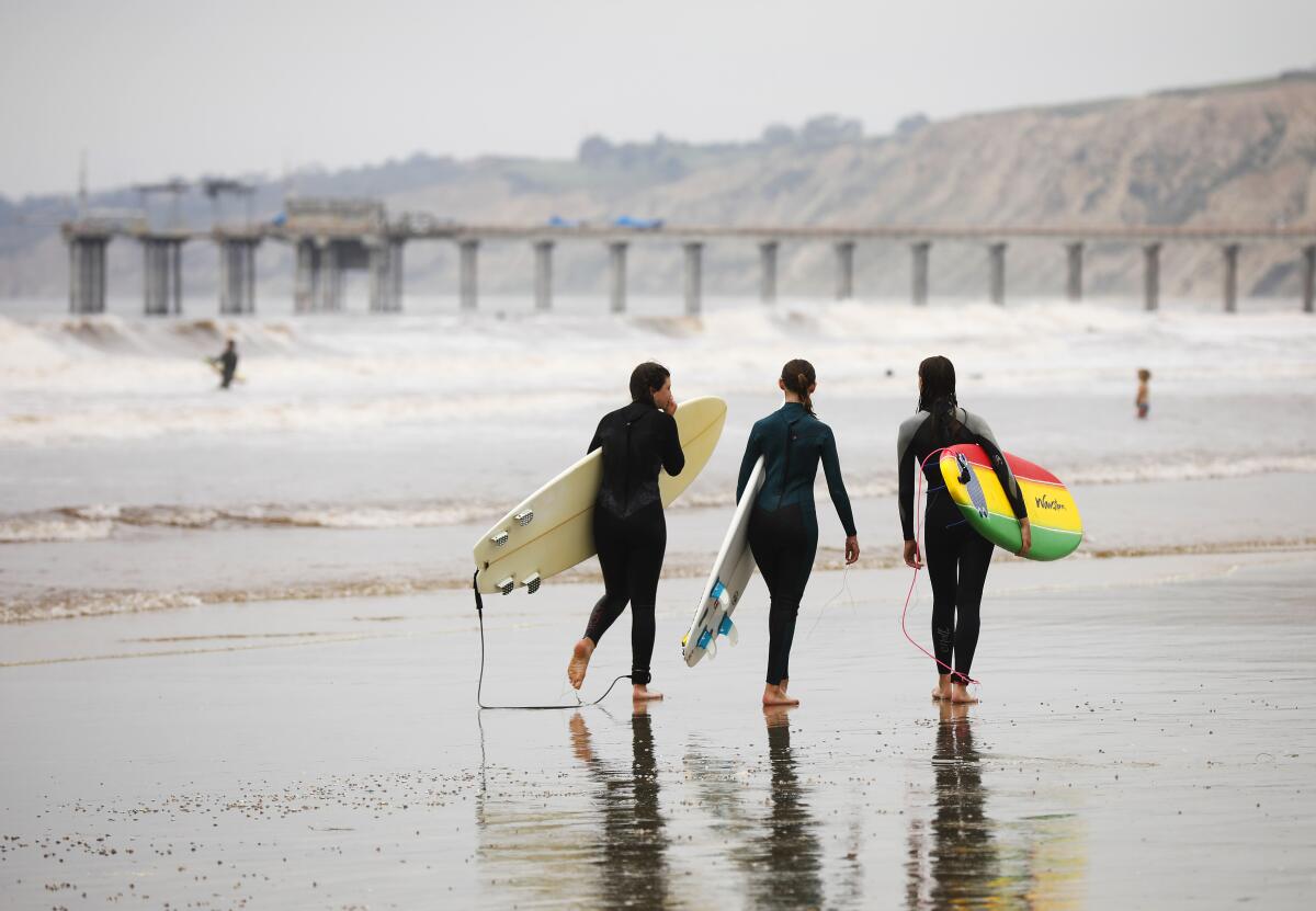 Three surfers in wetsuits carry surfboards on a beach