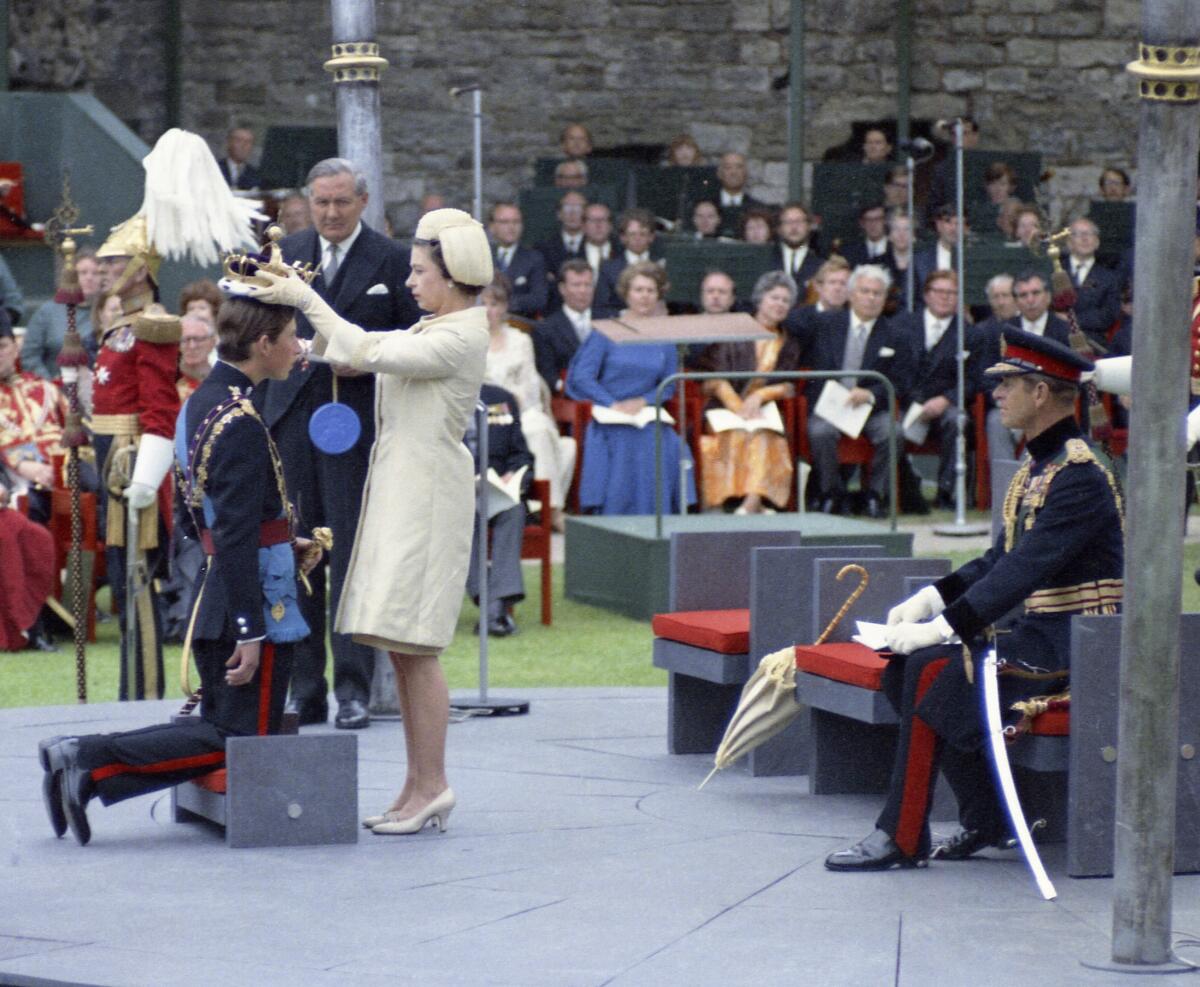 A woman in a cream-colored hat, dress and gloves, right, places a crown on a man in military uniform kneeling before her