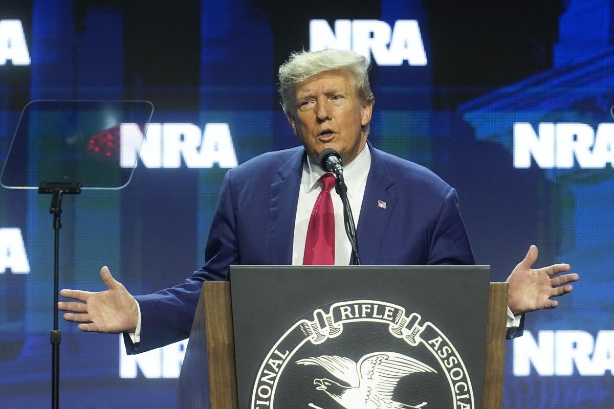 Trump speaks at a lectern with his hands out at his sides in front of a screen with NRA logos.