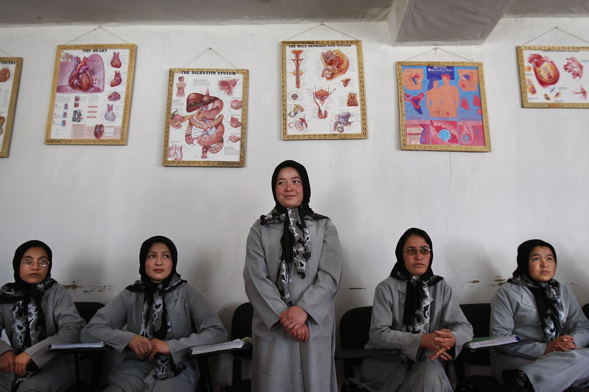 A young woman in a headscarf and gray uniform stands with four other women seated around her