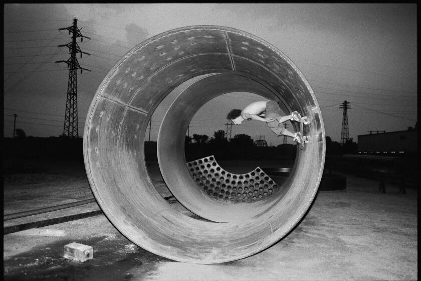 A black and white photo shows a skateboarder defying gravity as he skates the inside of a giant concrete water main pipe 