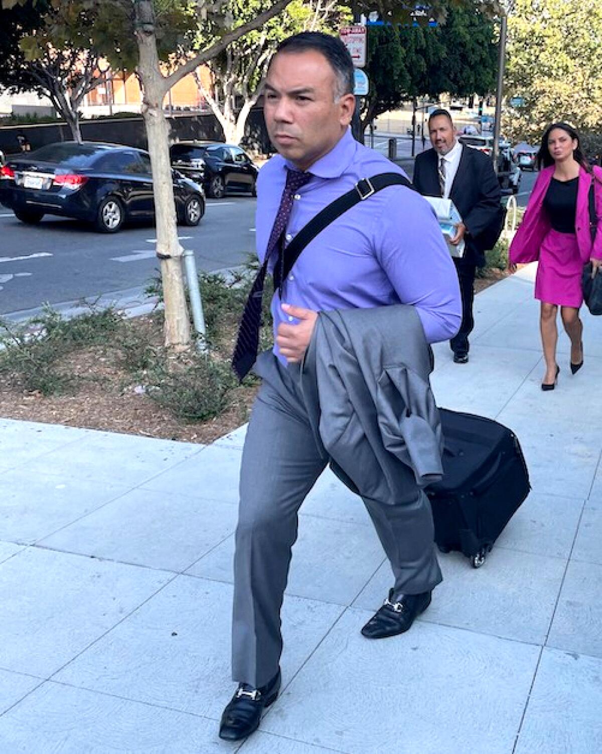 Zendejas Chavez with black and gray short hair in a lavender-colored dress shirt, wheeling a black bag on a city sidewalk