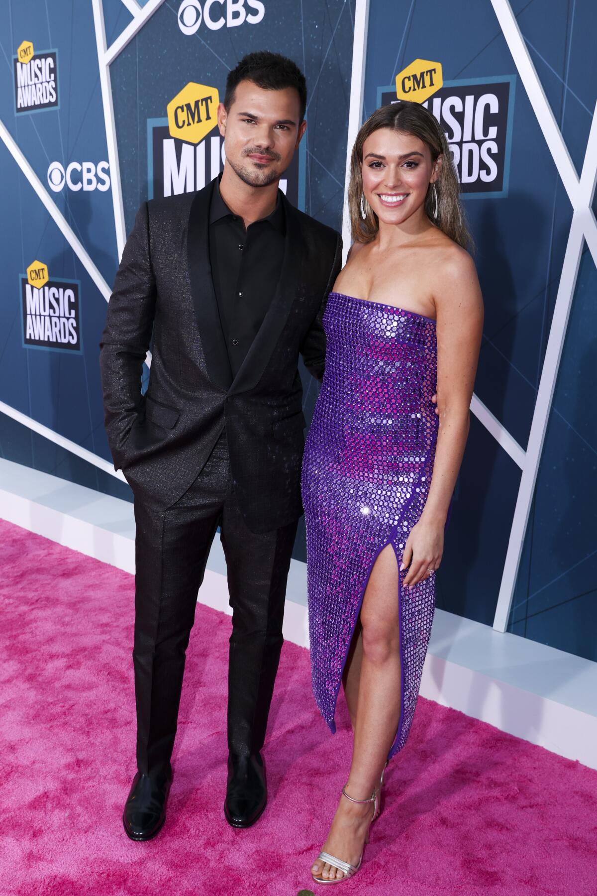 A young man wearing all black and a young woman wearing a sparkly strapless dress pose on a red carpet at an awards show