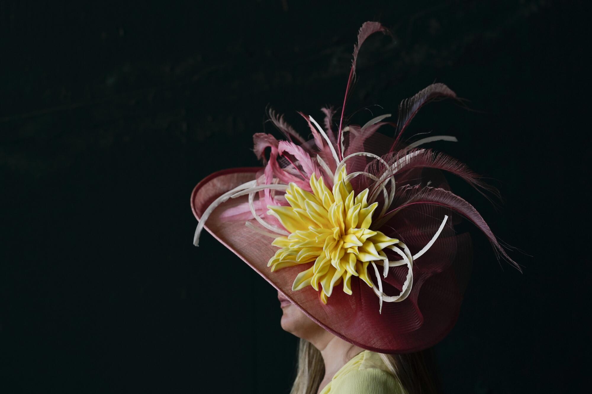 A woman wears a pink brimmed hat with a large yellow flower and pink feathers on it.