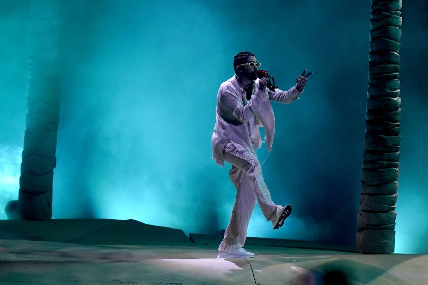 A man in sunglasses and a white suit performing on a stage filled with blue fog