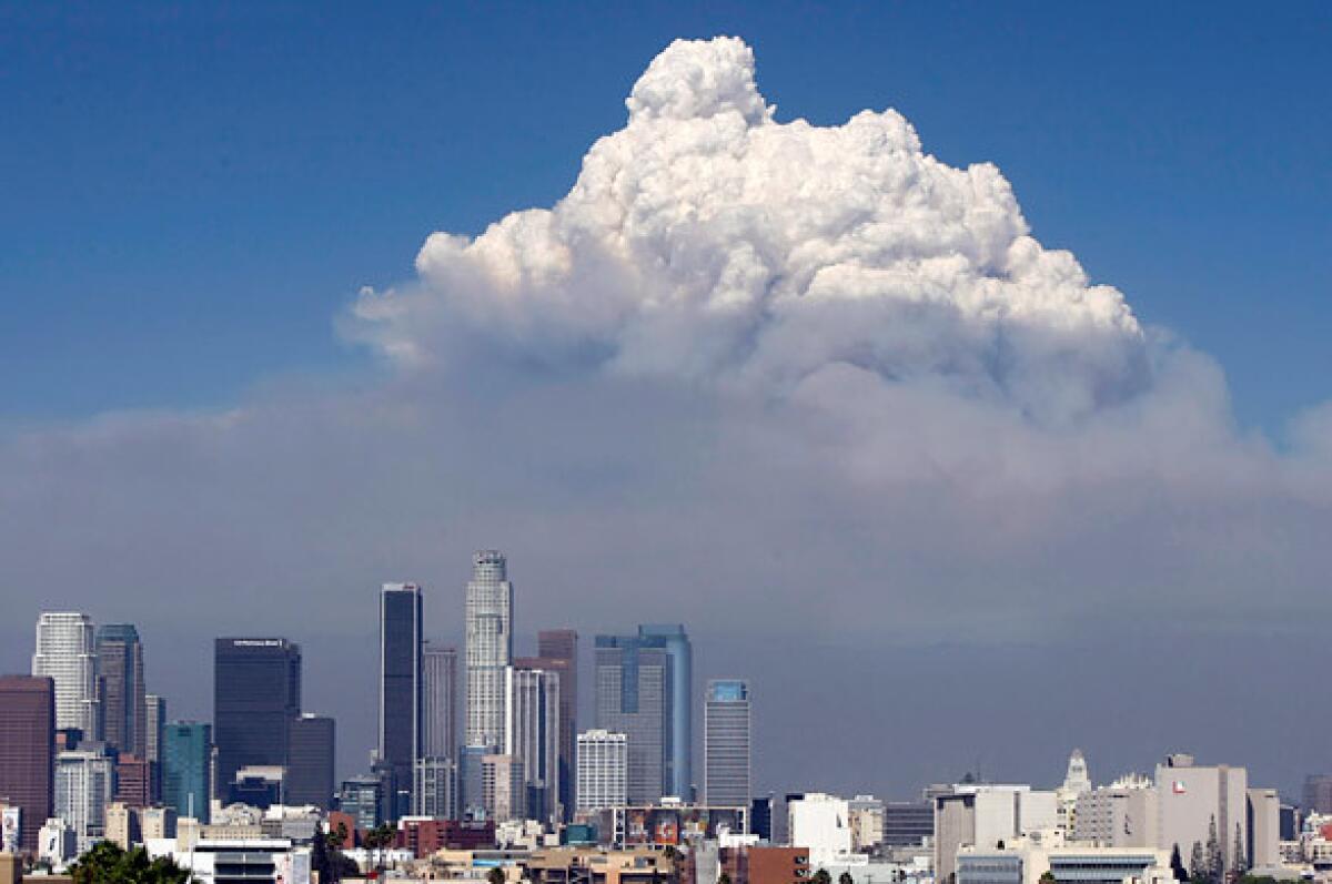 A large cloud with dark smoke rises in the background behind the downtown Los Angeles skyline