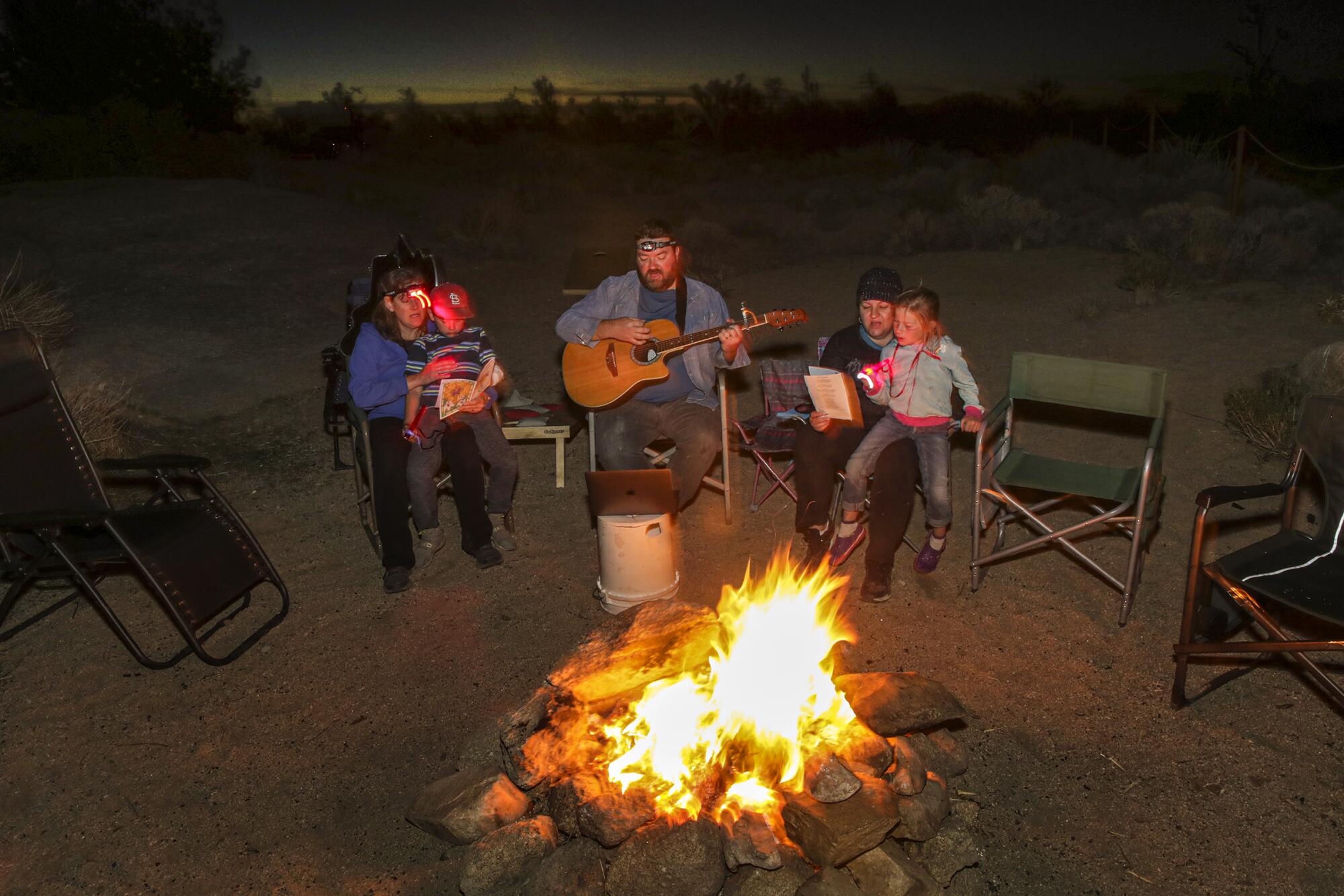 A man with a guitar and members of his family sit in folding chairs around a campfire at night