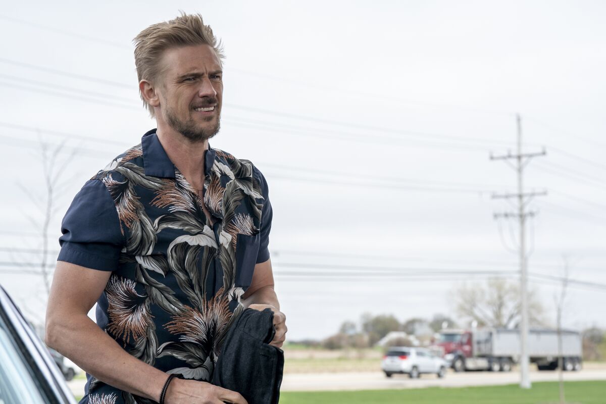 A man in a blue floral shirt stands with a jacket in his hands.