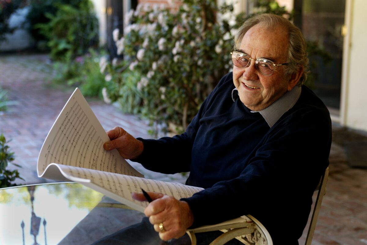 William Kraft, in a dark sweater, is seen holding sheet music while seated on an outdoor patio.