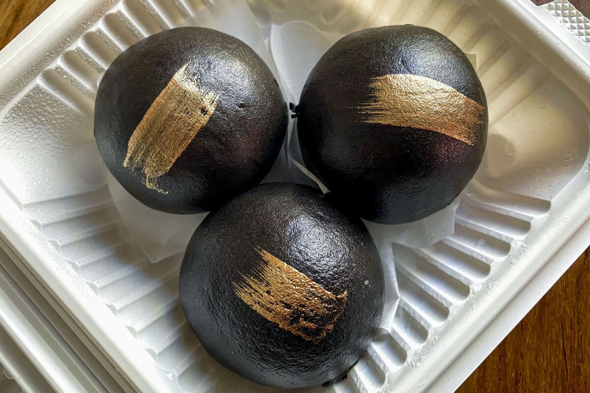 Three round black buns, each with a stripe of gold, in a takeout container
