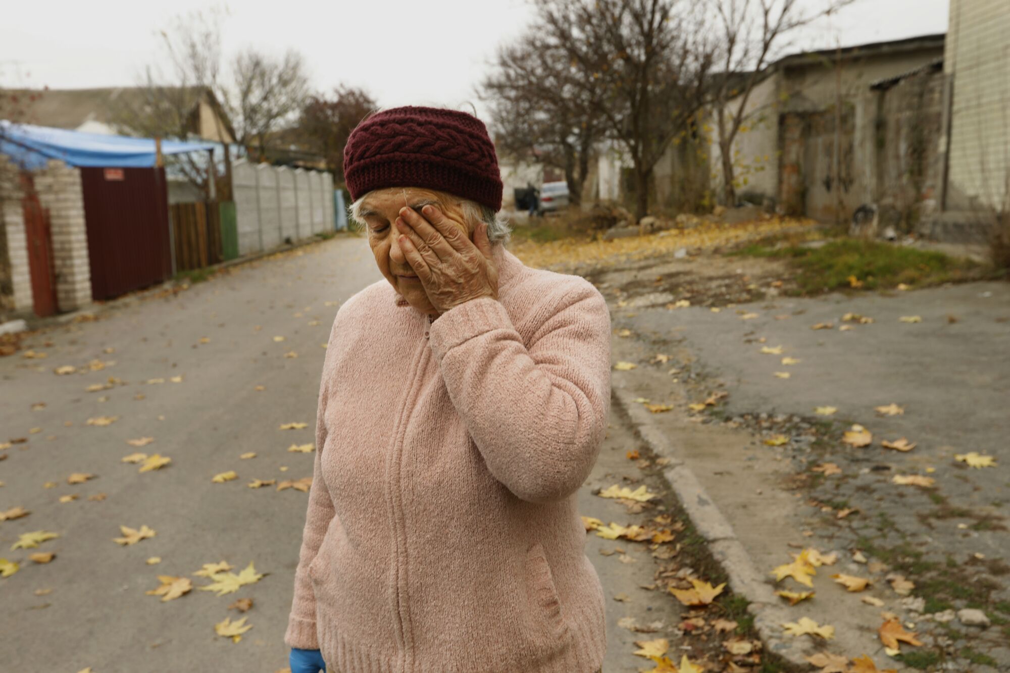 A woman in a pink cardigan and burgundy knit hat holds a hand to her face while standing on a street lined with buildings 